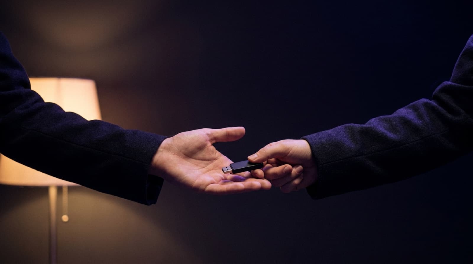 One person passing a small USB drive into another person's open hand in a dimly lit room